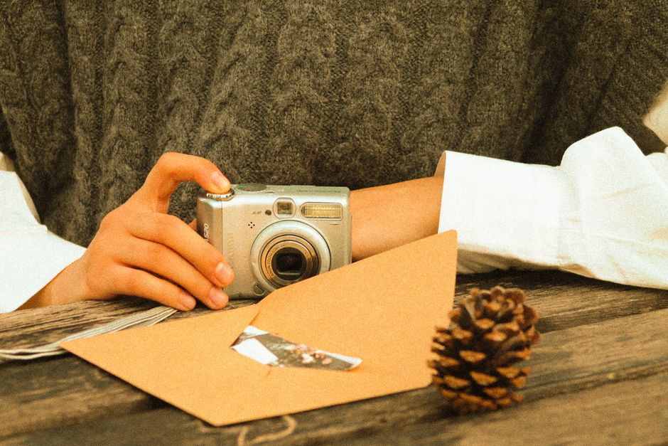 Close-up of hands holding a vintage camera with an envelope and pine cone on a rustic table.